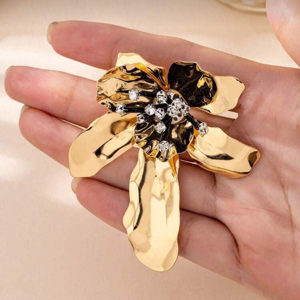 Brosa mare din metal auriu in forma de floare sculpturala cu petale lucioase, pistil negru si cristale albe tinuta in mana